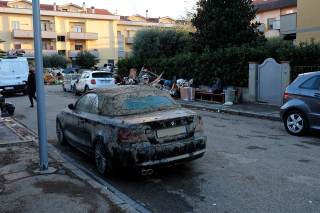 Macchina nera in primo piano, ricoperta di fango. Sullo sfondo mucchi di rifiuti fuori dalle case allagate.