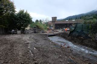 Fango, detriti lungo il percorso che ha preso il torrente verso la casa sullo sfondo. Mezzi al lavoro per il ripristino.