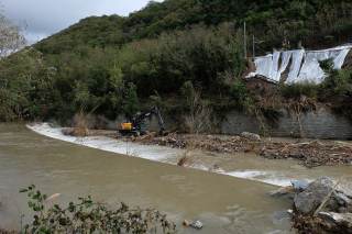 Ruspa che toglie detriti dal letto del fiume, sullo sfondo l\'area della ferrovia con degli smottamenti